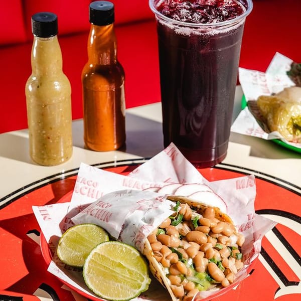 A bean taco with lime wedges, two hot sauce bottles, and a dark red drink on a red table.